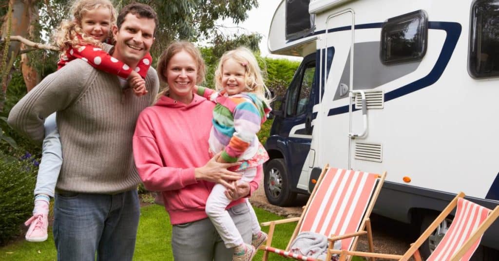 A family of four wearing colorful clothing smiles while standing next to a white RV, next to folding chairs.