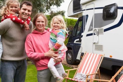 A family of four wearing colorful clothing smiles while standing next to a white RV, next to folding chairs.