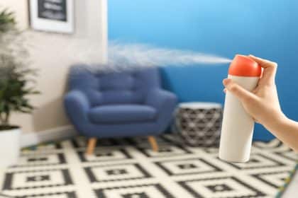 Close-up of a woman's hand spraying an air freshener into a room. It features a potted plant and a blue chair.