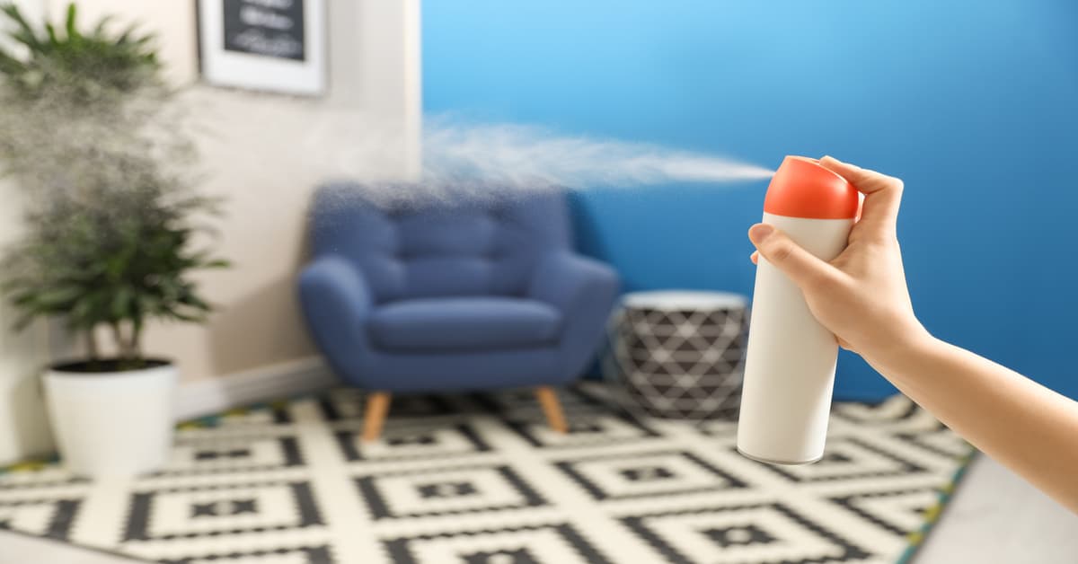 Close-up of a woman's hand spraying an air freshener into a room. It features a potted plant and a blue chair.