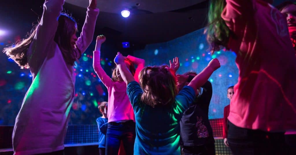 Several kids gather in a group, jumping around in a dimly lit room. A black light illuminates polka dots on the walls.