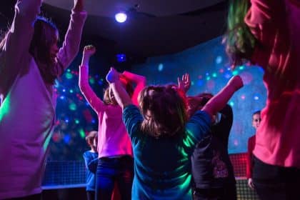 Several kids gather in a group, jumping around in a dimly lit room. A black light illuminates polka dots on the walls.