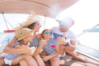 A mother, father, and two daughters laugh and smile together while wearing sunglasses on a sunny day aboard a boat.