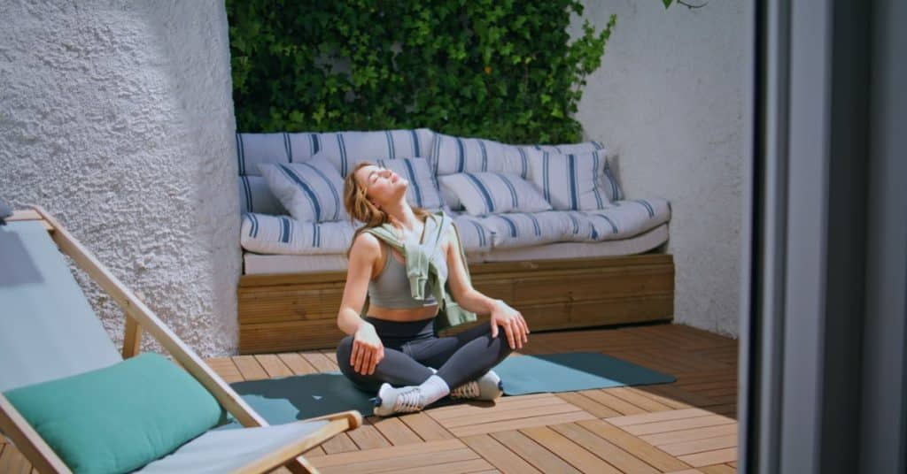 A young mom sits on her patio on a yoga mat in activewear as she gazes up at the sun for a moment of meditation.