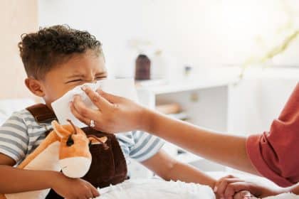 A mother uses a cloth to wipe at the nose of her child. The child is sitting in bed holding onto a stuffed animal.