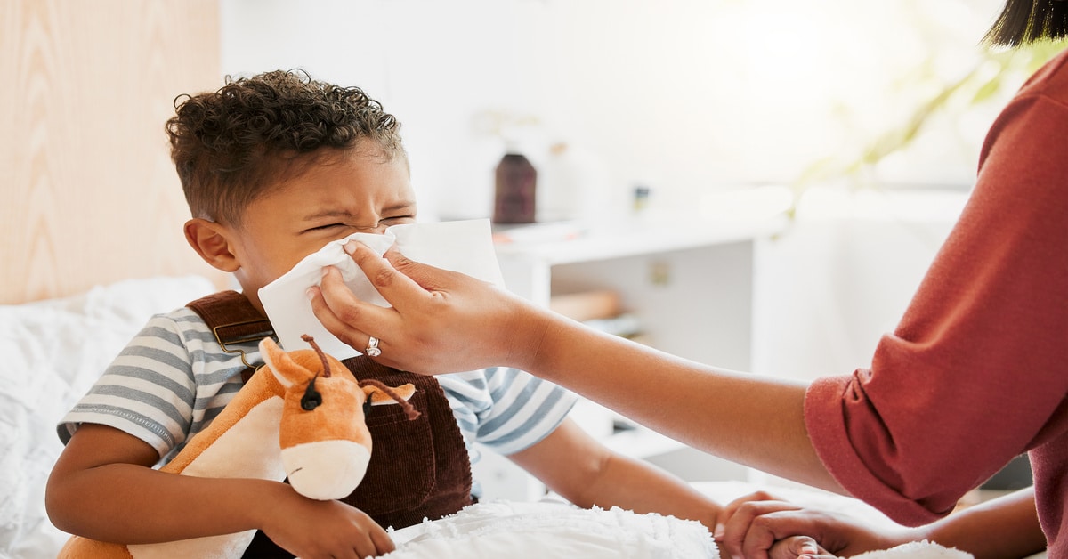 A mother uses a cloth to wipe at the nose of her child. The child is sitting in bed holding onto a stuffed animal.