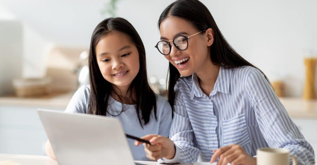 Happy mom helping her studying daughter, looking at the laptop screen together, sitting at the kitchen counter.