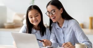 Happy mom helping her studying daughter, looking at the laptop screen together, sitting at the kitchen counter.