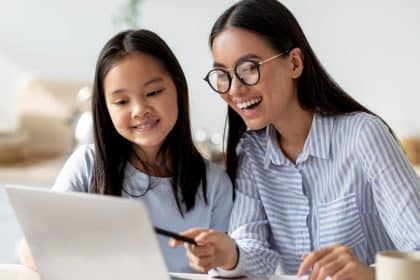 Happy mom helping her studying daughter, looking at the laptop screen together, sitting at the kitchen counter.