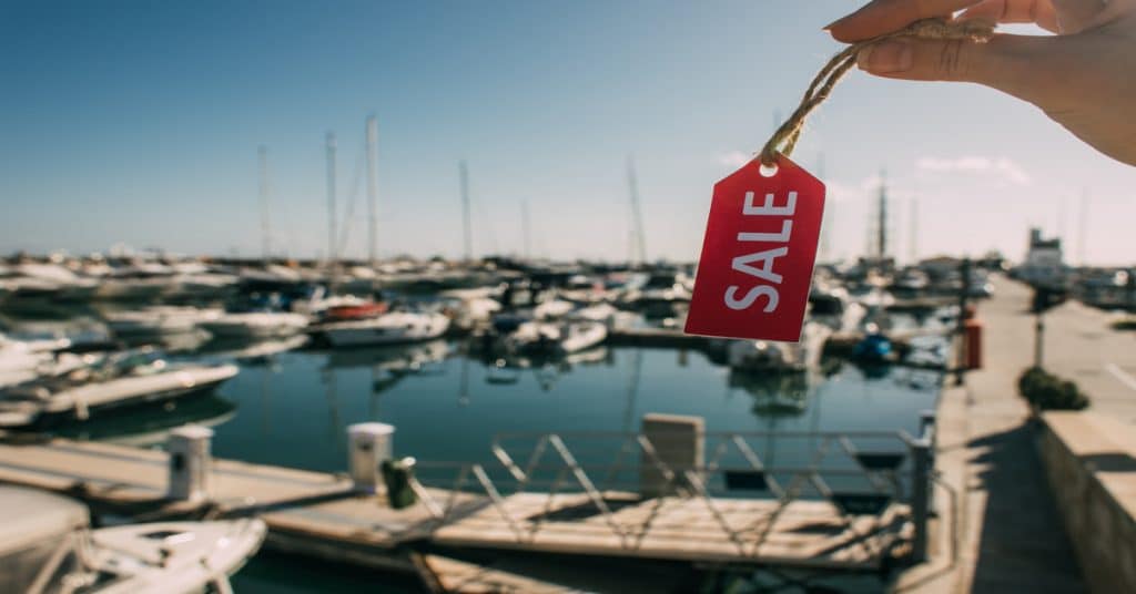 Someone holds up a small, red tag that reads "SALE." A blurred marina with boats in the water is behind the tag.