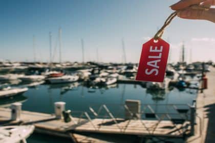 Someone holds up a small, red tag that reads "SALE." A blurred marina with boats in the water is behind the tag.
