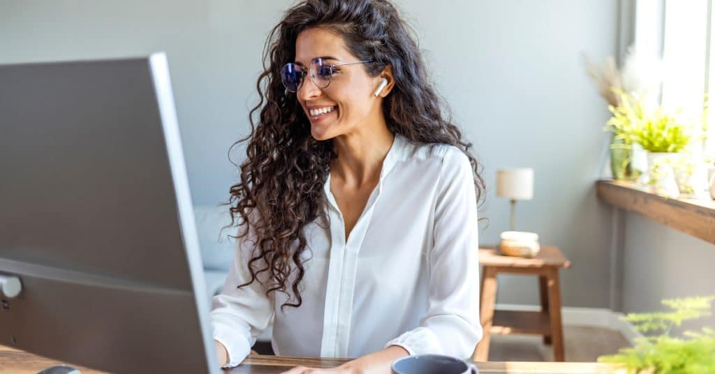 A smiling woman wearing glasses sits at a wooden desk, typing on a white keyboard and looking at the computer monitor.