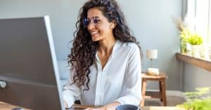 A smiling woman wearing glasses sits at a wooden desk, typing on a white keyboard and looking at the computer monitor.
