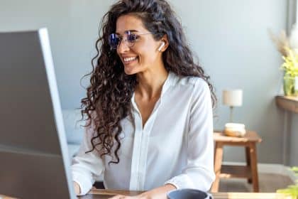 A smiling woman wearing glasses sits at a wooden desk, typing on a white keyboard and looking at the computer monitor.