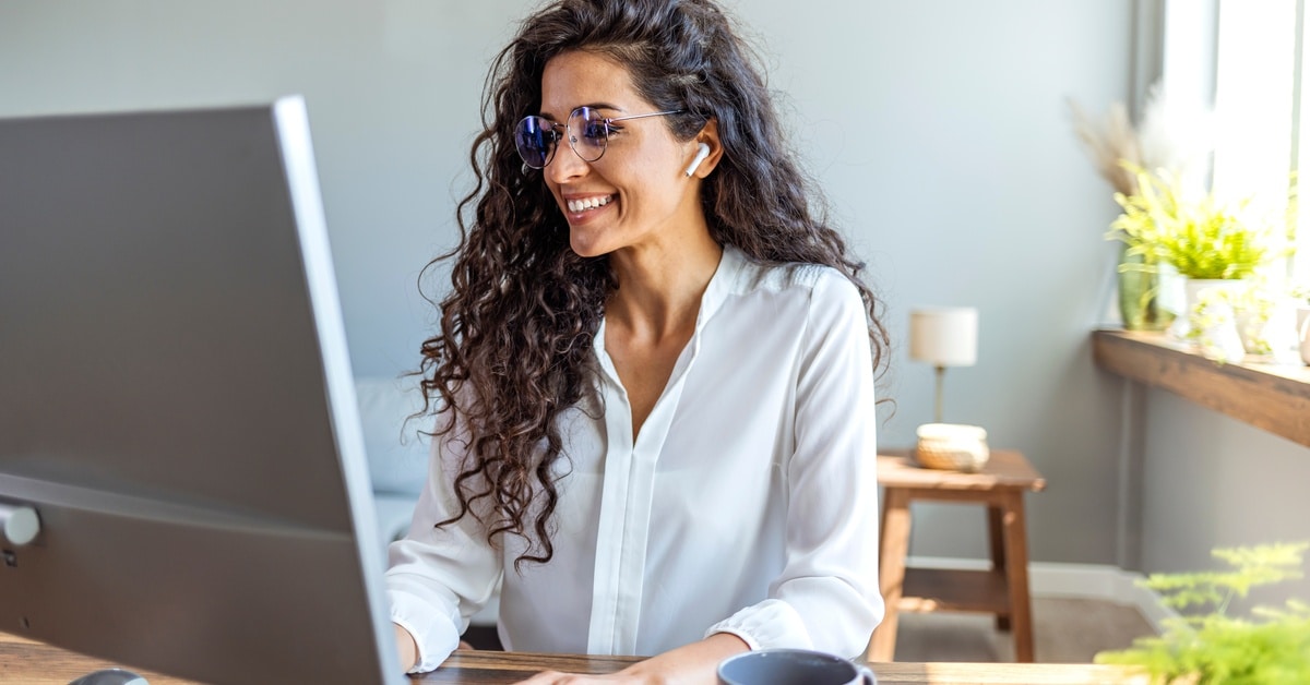 A smiling woman wearing glasses sits at a wooden desk, typing on a white keyboard and looking at the computer monitor.