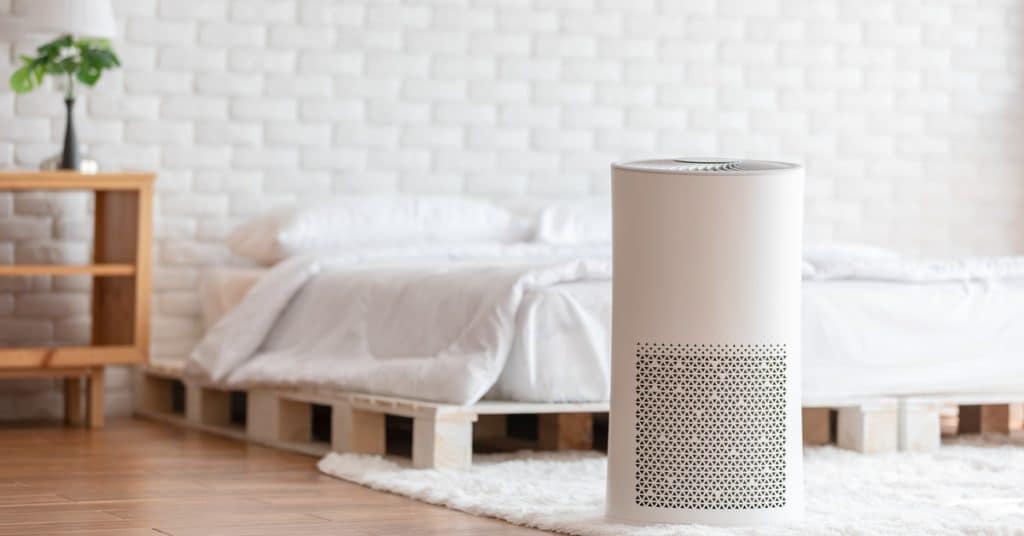 A white air purifier sits on a white rug in front of a bed that has white sheets. A white wall is behind the bed.