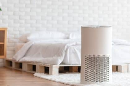 A white air purifier sits on a white rug in front of a bed that has white sheets. A white wall is behind the bed.