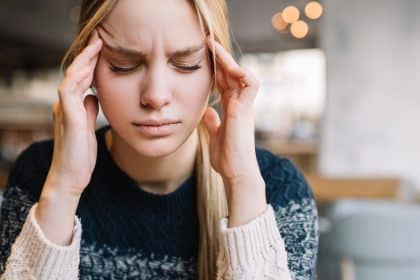A close-up view shows a young woman closing her eyes and massaging the side of her temple with her hands.