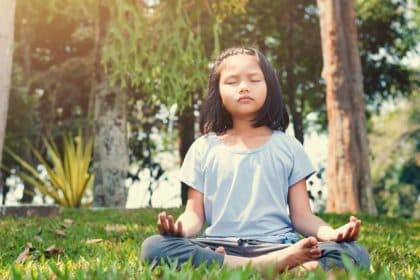 A child sits cross-legged on grass with eyes closed and hands resting on knees while positioned outdoors in sunlight.