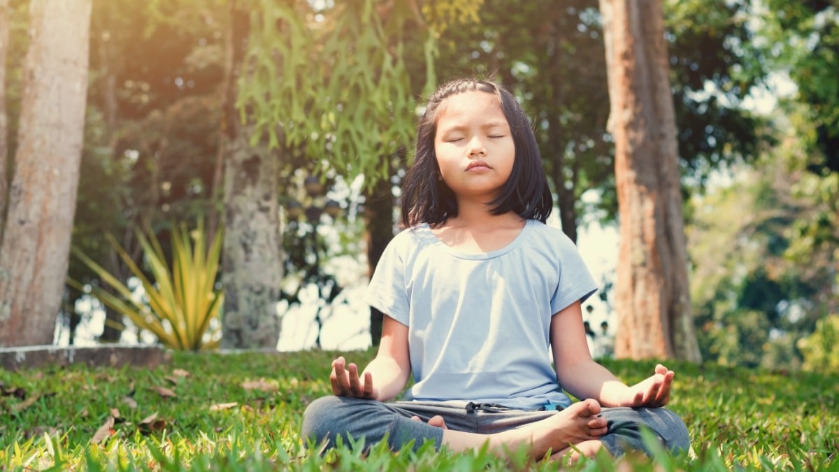 A child sits cross-legged on grass with eyes closed and hands resting on knees while positioned outdoors in sunlight.