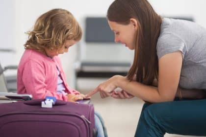 A woman kneels and speaks to a child sitting on a suitcase in an airport waiting area with empty seats nearby.