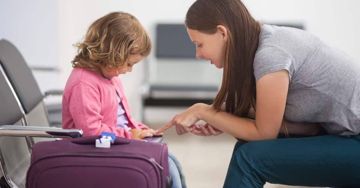 A woman kneels and speaks to a child sitting on a suitcase in an airport waiting area with empty seats nearby.