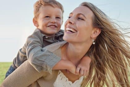 A smiling mother carries her smiling son on her back in a sunny field as the wind blows through her hair.