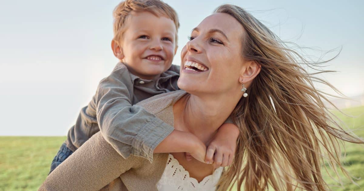 A smiling mother carries her smiling son on her back in a sunny field as the wind blows through her hair.