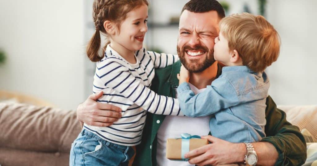 A happy, smiling father with two kids hugging either side of him on a couch. The son kisses him on the cheek.
