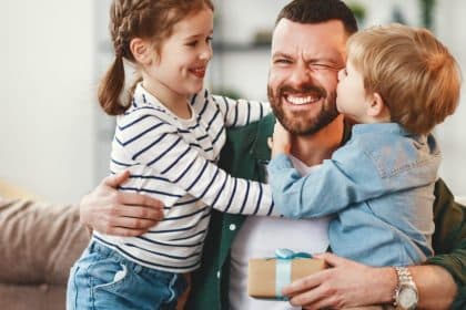 A happy, smiling father with two kids hugging either side of him on a couch. The son kisses him on the cheek.