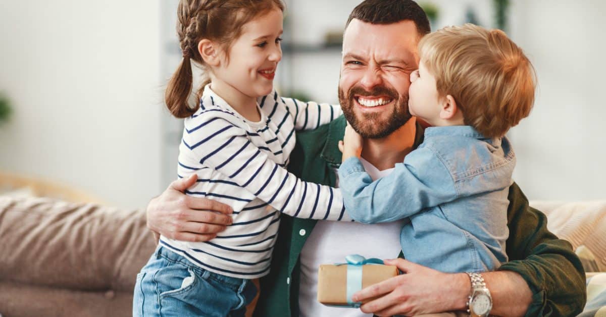 A happy, smiling father with two kids hugging either side of him on a couch. The son kisses him on the cheek.
