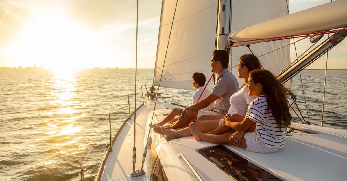 Family sitting on sailboat at sunset, looking toward horizon over calm sea, enjoying warm light and peaceful atmosphere.