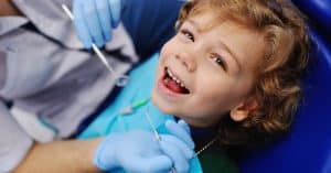 A young boy lies back in a dental chair while wearing a blue bib. He smiles as the dentist holds up two tools.