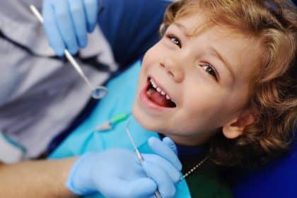 A young boy lies back in a dental chair while wearing a blue bib. He smiles as the dentist holds up two tools.
