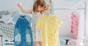 A child standing near a clothing rack and looking at dresses hanging in a bright children's room with white walls.