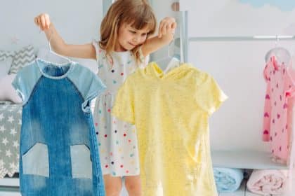 A child standing near a clothing rack and looking at dresses hanging in a bright children's room with white walls.