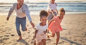 A family with two adults and two children running across a sandy beach beside ocean waves at sunset on a clear evening.