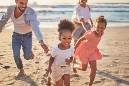 A family with two adults and two children running across a sandy beach beside ocean waves at sunset on a clear evening.