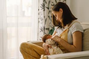 A mother sits in a tan chair breastfeeding her newborn baby. She is looking at him lovingly and holding his hand.