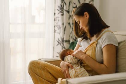 A mother sits in a tan chair breastfeeding her newborn baby. She is looking at him lovingly and holding his hand.