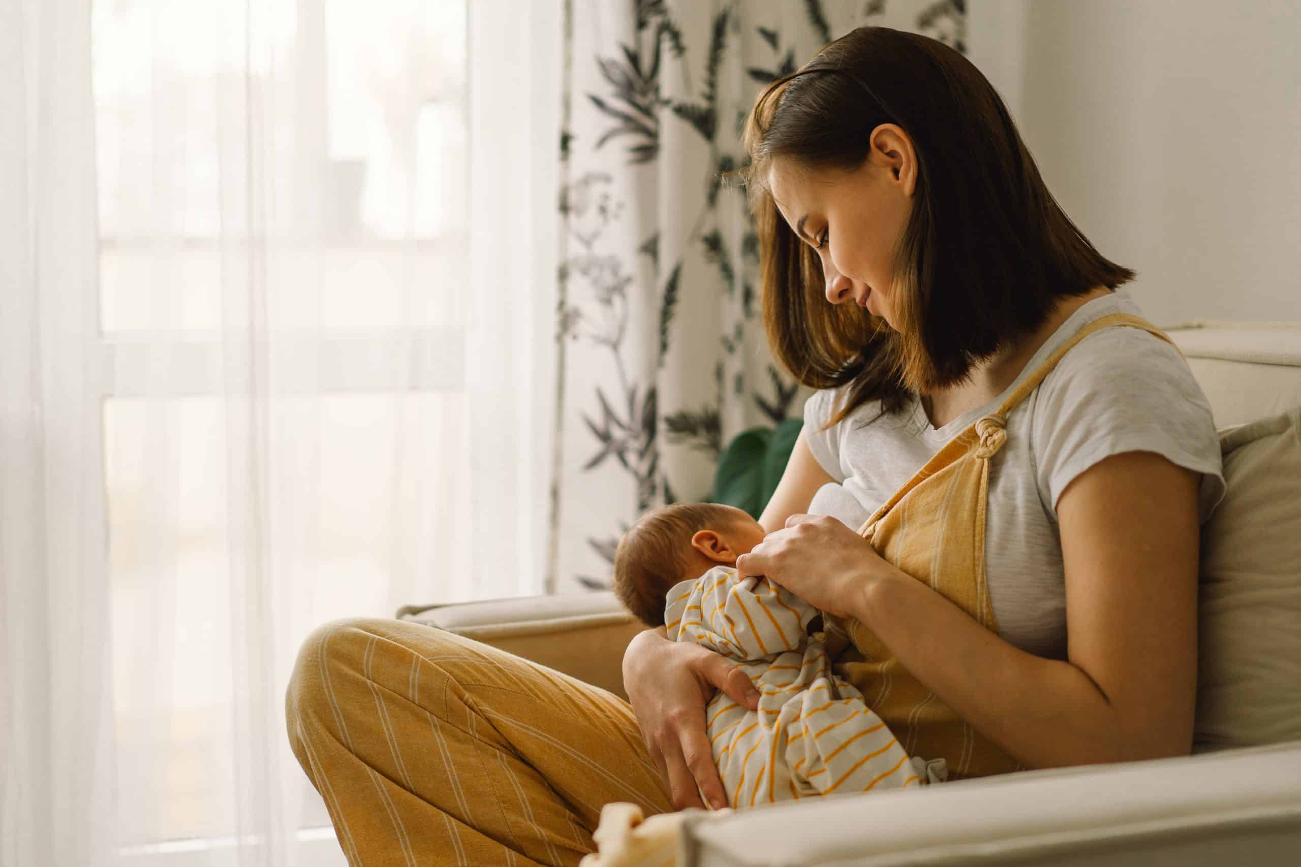 A mother sits in a tan chair breastfeeding her newborn baby. She is looking at him lovingly and holding his hand.