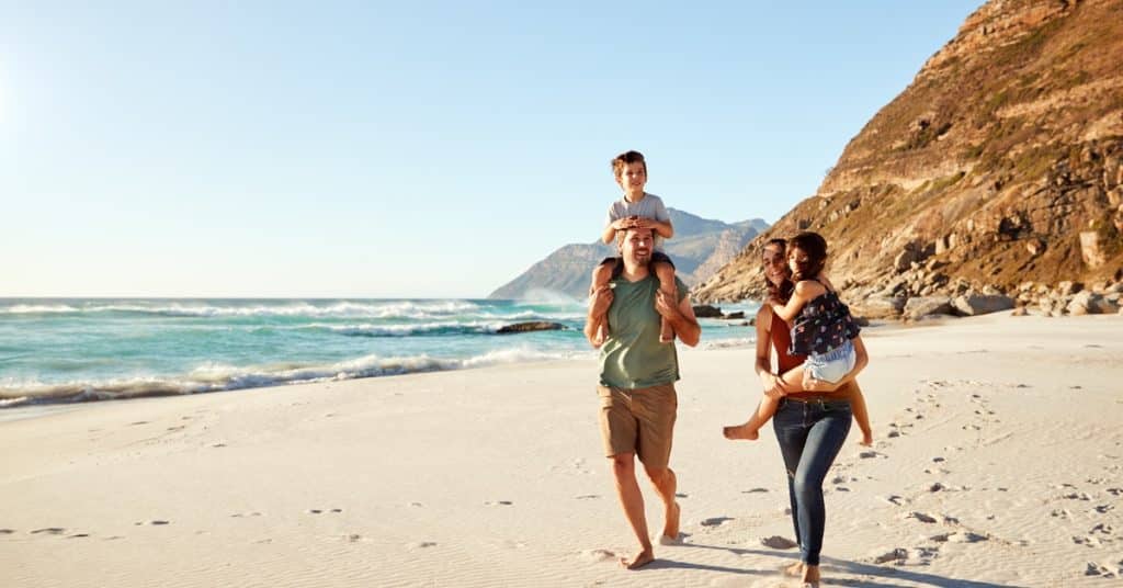  Family walks along sandy beach by ocean, parents carrying children with rocky hillside and waves in background.