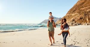  Family walks along sandy beach by ocean, parents carrying children with rocky hillside and waves in background.