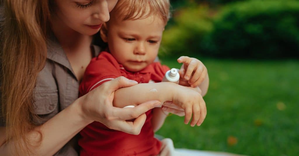 A woman applying cream to a child’s hand outdoors, with several bushes and grass visible in the background.