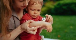 A woman applying cream to a child’s hand outdoors, with several bushes and grass visible in the background.