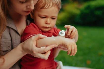 A woman applying cream to a child’s hand outdoors, with several bushes and grass visible in the background.