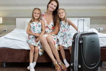 A woman sits on the edge of a hotel bed between two twin girls. They all wear dresses. A rolling suitcase is near them.