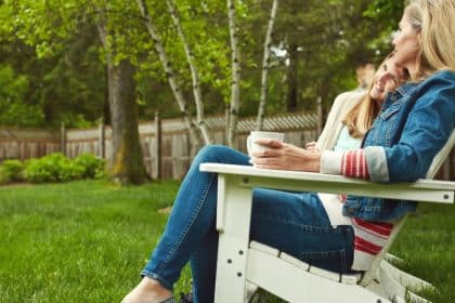 Two women sitting together in white lawn chairs on green grass beside trees and a wooden fence.