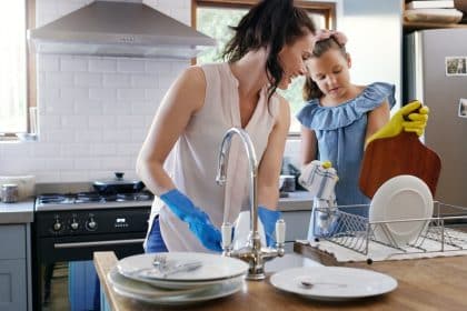A mom and daughter wearing rubber gloves cleaning dishes in a residential kitchen during the daytime.