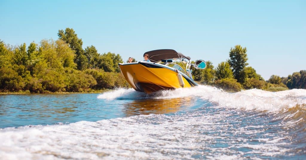 A yellow speedboat cuts across a lake at high speed, creating a foamy wake as passengers enjoy a sunny day on the water.